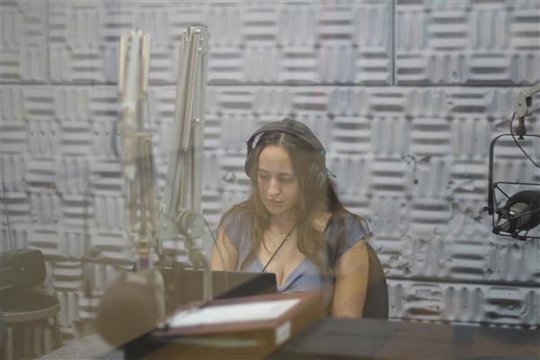 A student sits behind a radio mixing board. She is wearing headphones and looking down at the board. The photo is taken from outside the studio, through the window glass. Even though you can't hear what she's playing, her serious expression lets you know that she is doing a great job.