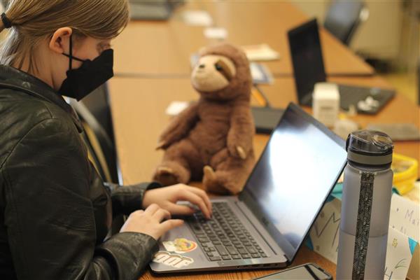 A student is typing on a laptop computer. To the left of the computer is a stuffed animal sloth. To the right of the computer is a water bottle. The student is planning an excellent radio program on KECG.
